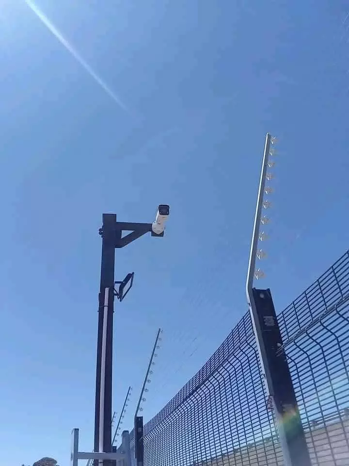 Security camera on a tall post and a wire security fence against a blue sky